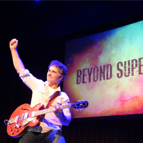 A man, Noel Fitzpatrick, is posing with his electric guitar. In front of a screen displaying, "BEYOND SUPERVET", the name of his tour.