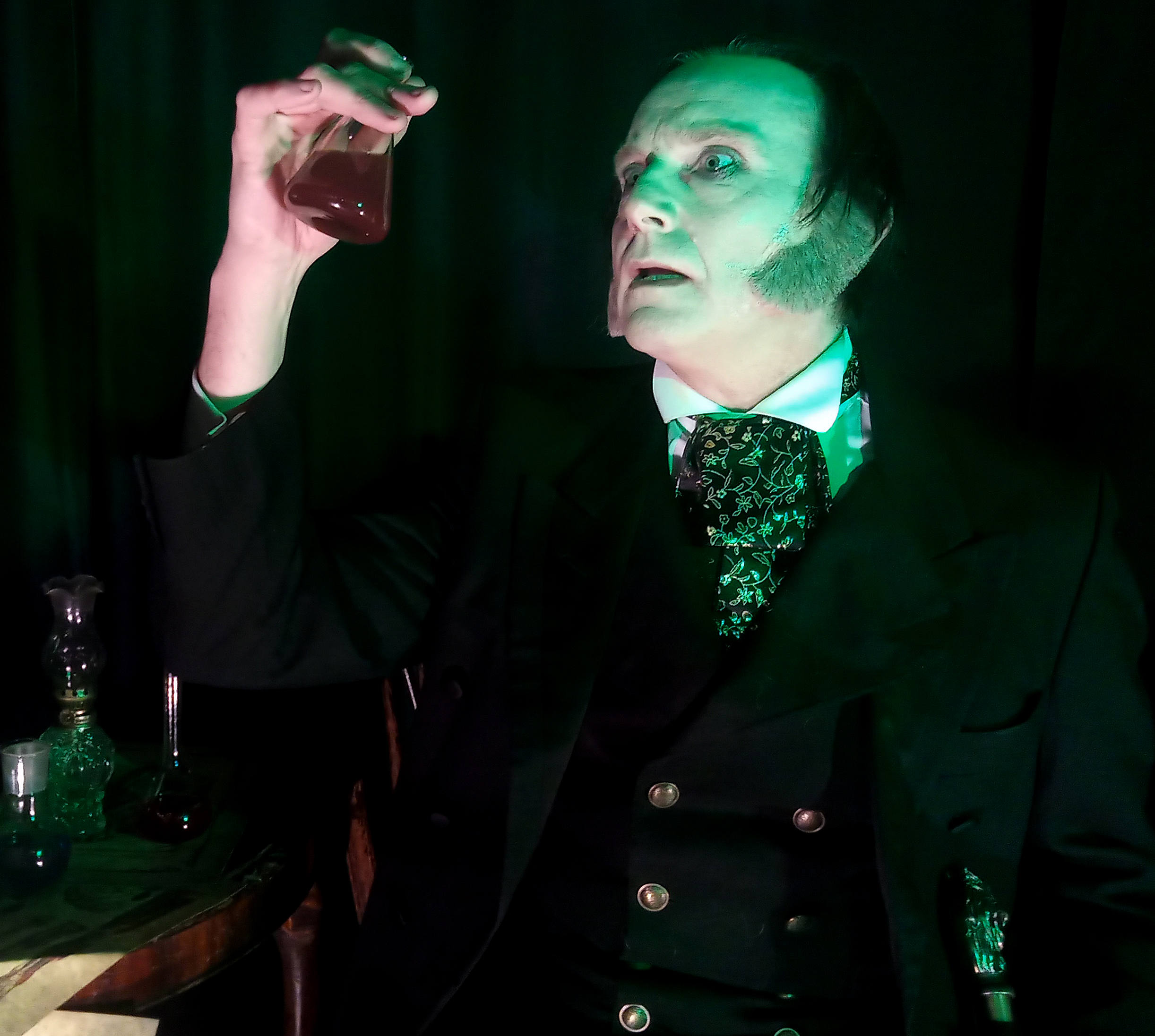 A pale faced man in Victorian dress holds up a glass vial with red liquid in it