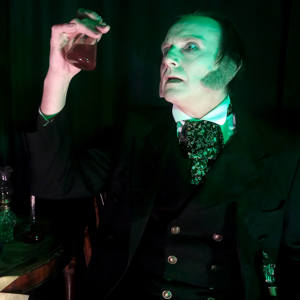 A pale faced man in Victorian dress holds up a glass vial with red liquid in it