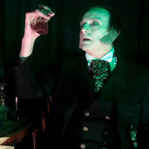 A pale faced man in Victorian dress holds up a glass vial with red liquid in it