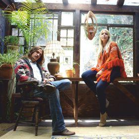 A man is sat on a chair and a woman is sat on a table in a rustic, wooden room. 