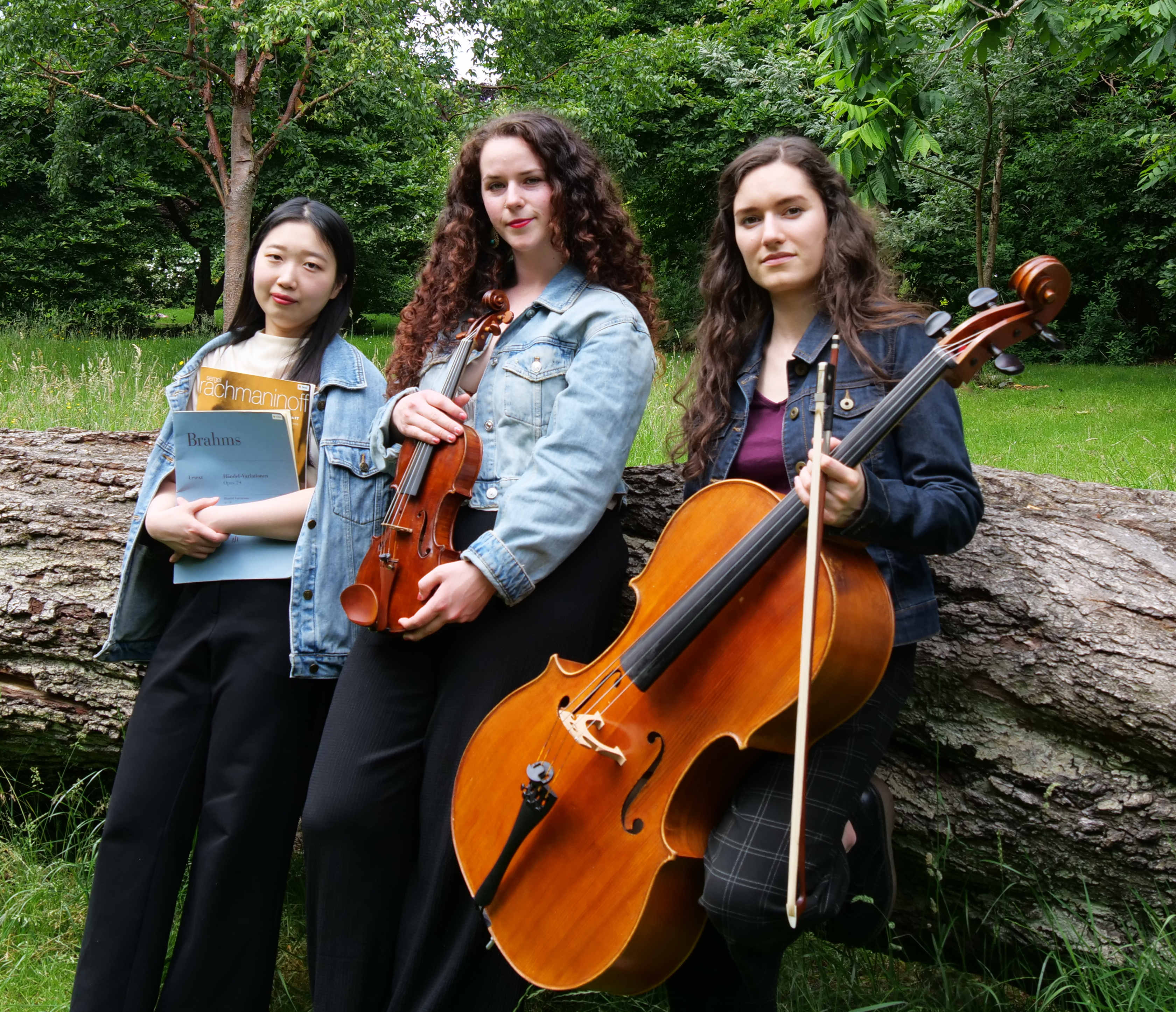 Three musicians leaning against a horizontal tree trunk holding music, a violin and a cello
