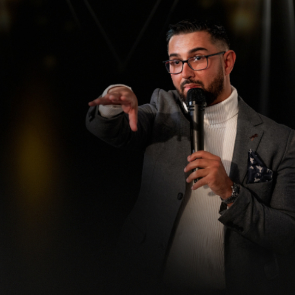 Calvin Price Medium wearing a cream turtle neck jumper and grey jacket, speaking into a microphone